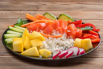 Top view of a Poke salad from hawaii in black plate on wooden background. Close up. Soft focus. To prepare it just take rice, salmon and any vegetables.