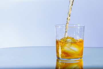 Whiskey pours into a glass of ice and whirls around, against a neutral blue background