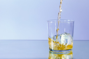 Whiskey pours into a glass of ice and spills, against a neutral blue background