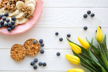 Served white table, a plate of banana pieces, blueberry and oatmeal cookies. Yellow tupic flowers decorations. 