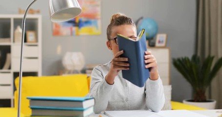 Pretty Caucasian teen small schoolgirl in glasses sitting at the desk in her room, opening a book and reading a textbook while doing homework. - Powered by Adobe