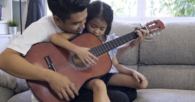 Father teach children guitar. Asian man and child sitting together on sofa in living room learning to play guitar. 
