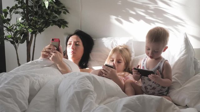 Young mother, daughter and little son are using gadgets lying on a white bed. Concept modern family during weekend at home. Contemporary technology.