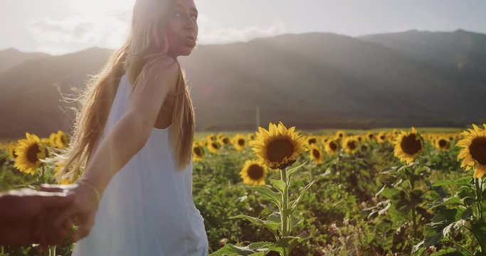 Young couple holding hands walking through sunflower field at sunset in slow motion, beautiful young woman looking back at her boyfriend and smiling