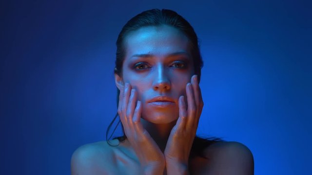 Shiny futuristic fashion model in cold blue neon lights touching and covering her face with hands on blue background.