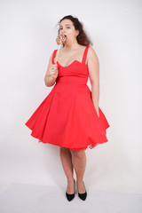 pretty chubby fashion girl wearing red pinup dress and posing with tasty colorful lollipop on white studio background