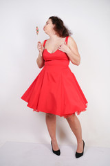 pretty chubby fashion girl wearing red pinup dress and posing with tasty colorful lollipop on white studio background