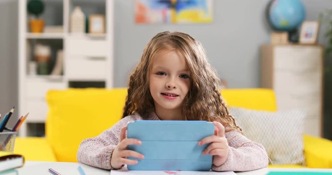 Portrait of the cute and pretty Caucasian little girl smiling to the camera happily while sitting at the desk and watching something on the tablet device in her room. Close up.