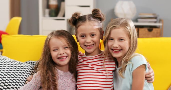 Portrait shot of the beautiful Caucasian three small girls sisters sitting on the yellow couch in the living room and smiling to the camera cheerfully. Close up.