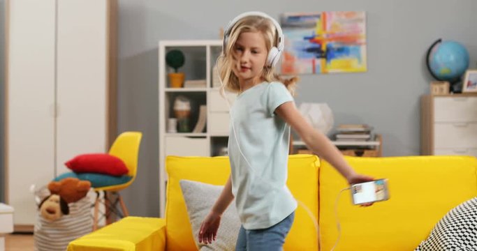 Cute Caucasian pretty blonde girl in headphones dancing and jumping while listening to the music on the smartphone player in the cozy living room.