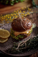 Dark burger with grain bread on dark ceramic plate, salad, rosemary and lemon