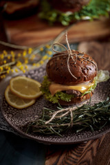 Dark burger with grain bread on dark ceramic plate, salad, rosemary and lemon