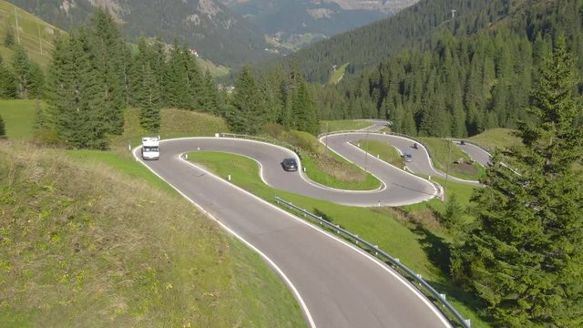 AERIAL: Flying over a recreational vehicle driving through corners on a bendy Italian road, followed by other cars and motorcycles. Idyllic small village is seen through the trees and Italian Alps.