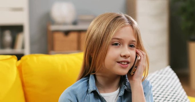 Caucasian pretty teen blond girl resting on the yellow couch in the living room and talking on the phone. Close up.
