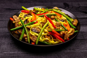 Appetizing stir fried noodles with garlic sprouts, mushrooms, red pepper and chicken meat on a black rustic background of a wooden table. A popular Asian dish