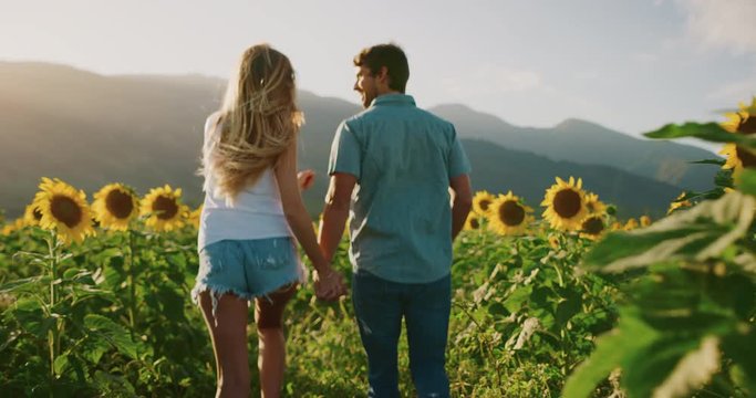 Young attractive happy couple holding hands and running joyfully together through sunflower fields in slow motion at sunset, summer romance