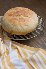 Round Loaf of Freshly Baked Sourdough Bread on Cooling Rack on Wood Table with Yellow and White Kitchen Towel