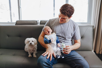 Young Caucasian father feeding newborn baby with milk. Male man parent with dog and yawning child...