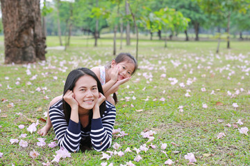 Fototapeta premium Happy little girl riding on back her mom lying on green field with fully fall pink flower in the garden outdoor. Happy loving family.
