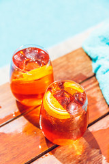 Two glasses of aperol spritz and negroni cold cocktail on wooden edge of swimming pool with...