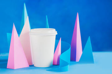Modern coffee header. Blank paper cup on an abstract background with modern paper sculpture. Paper pyramids and curves design in pastel colors.