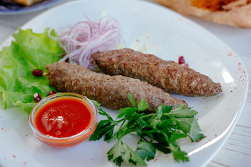 Meat kebabs on white plate with green herbs and red sauce.