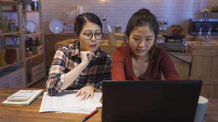 two female roommates checking their bills at home kitchen wooden table. girls accounting on laptop computer online on internet bank website. young women friends living together lifestyle concept.
