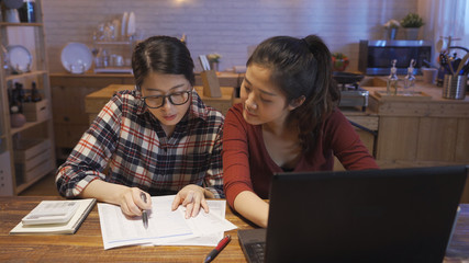 two asian women managing finances reviewing bank accounts using laptop computer and calculator at modern kitchen wooden table. sisters doing paperwork together paying taxes online on notebook pc