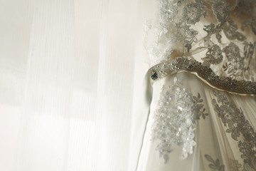 Beautiful white wedding dress hanging near a window in hotel room, morning wedding preparation, white lace dress for bride on hanger close-up