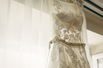 Fototapeta na wymiar Beautiful white wedding dress hanging near a window in hotel room, morning wedding preparation, white lace dress for bride on hanger close-up