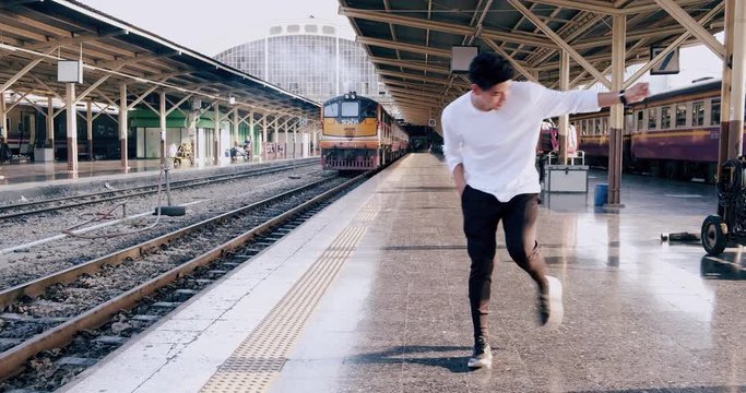 Wide shot of young hipster doing freestyle street dancing in train station while waiting for train. Asian man.