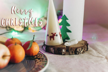 merry christmas text sign on candle with reindeers and christmas tree on rustic  table with fruits and  colorful lights on background. space for text. seasonal holidays greetings