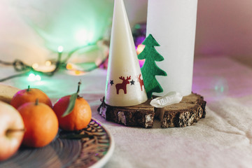 candle with reindeers and christmas tree on rustic christmas table with fruits and  colorful lights on background. space for text. seasonal holidays greetings