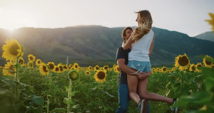 Young attractive couple romantically playing in sunflower fields at sunset in slow motion, young attractive man twirling and embracing woman
