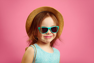 Portrait of beautiful little girl with straw hat and sunglasses, wears blue dress, stands on pink...