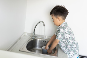 The boy is helping the mother to do the housework by washing a glass and a dish.