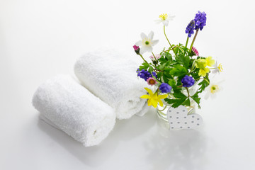 handtücher neben einer Cremedose vor weißem Hintergrund und Blumen in einer Vase.