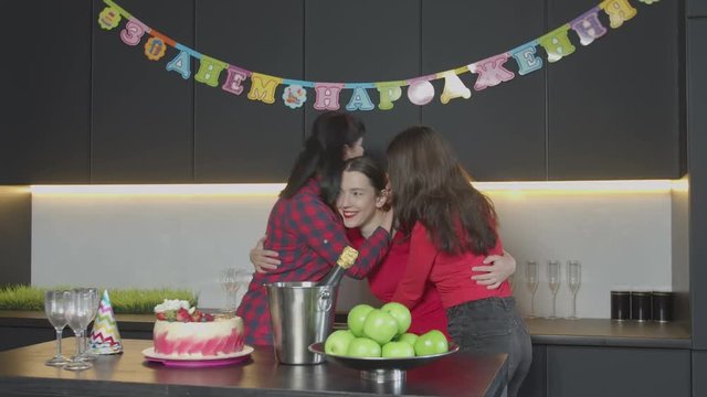 Happy carefree birthday girl enjoying embrace and kisses of mother and best friend during celebration of thirtieth anniversary in narrow circle. Pretty woman accepting congratulations in home kitchen.