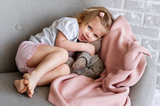 Little charming girl sits on a gray sofa. She plays with a gray toy bunny. They are covering with knitted pink plaid. Behind the sofa we see a brick gray wall.