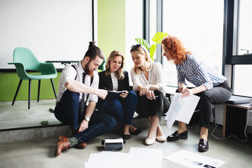 A group of young business people sitting on the floor in an office, talking.