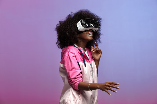 Mod young brown-haired curly girl dressed in the pink sports jacket uses the virtual reality glasses in the studio on neon background