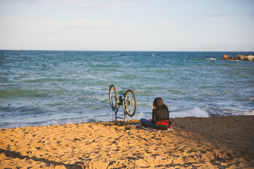 Girl with a bike is sitting on the beach. Hipster near the sea. Cycling hobby. Summer time. Travel concept.