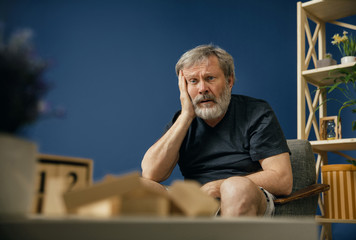 Acute pain and madness. Old bearded man with alzheimer desease sitting on the chair and suffering...