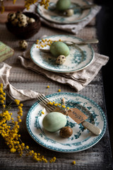 Orthodox Easter table setting with three vintage plates with green ornament, forks, dyed green eggs, nest with quail eggs, yellow flowers
