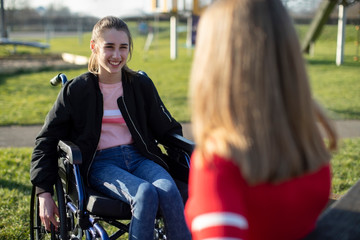 Fototapeta na wymiar Teenage Girl In Wheelchair Talking With Friend In Park