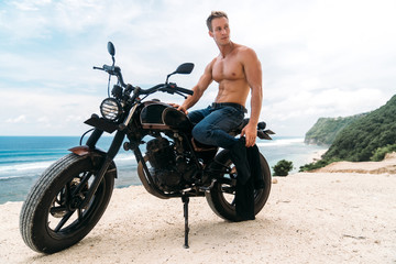 Obraz na płótnie Canvas Sexy athletic man with perfect naked body sitting on motorbike, ocean waves and beautiful mountains on background