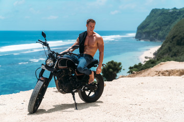 Fototapeta na wymiar Sexy athletic man with perfect naked body sitting on motorbike, ocean waves and beautiful mountains on background
