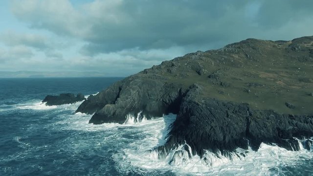Aerial dolly in towards a rocky irish island with waves crashing upson the shores with rough water all around on our a cloudy cold day