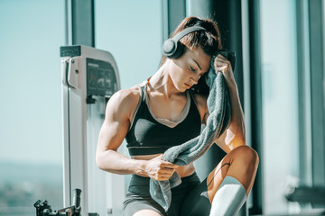 Serious muscular strong female bodybuilder with ponytail and headphones wiping sweat while sitting...