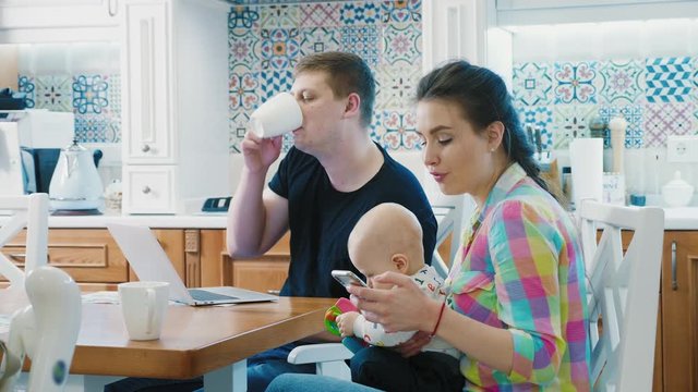 Dad entertains the kid with a toy. Young mom holds her child in a bright kitchen interior, embraces little son, talking on the smartphone, uses electronic devices.